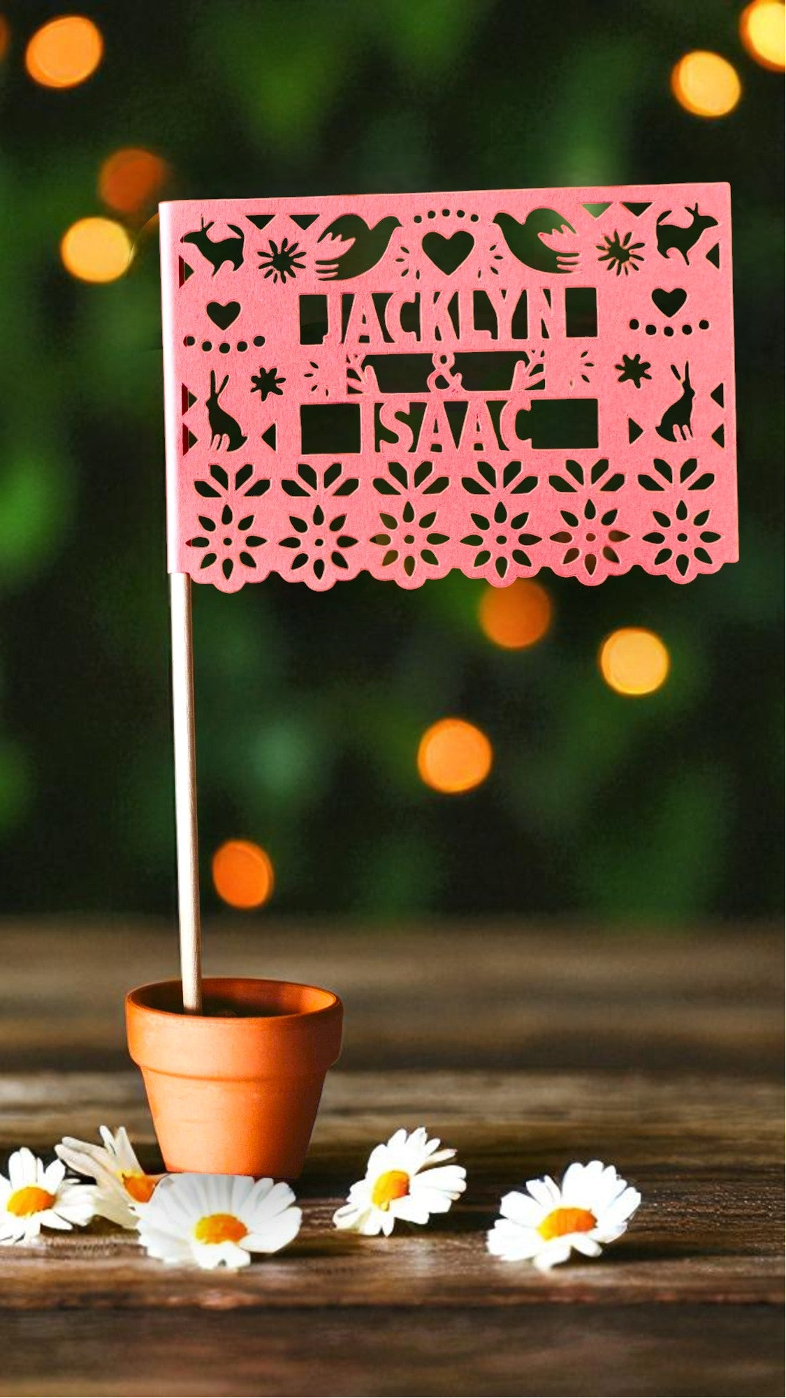 A pink papel picado table flag with the name 'MACKENZIE ISAAC' stands in a small terracotta pot on a wooden surface, next to a potted succulent and white daisy flowers, with a blurred green bokeh background.